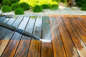 A close-up of a power washer from a top pressure washing service in Cabell County, WV, cleaning a wooden deck. The washer sprays water, creating a stark contrast between cleaned and uncleaned areas. Lush green bushes and decorative white stones provide a vibrant backdrop.