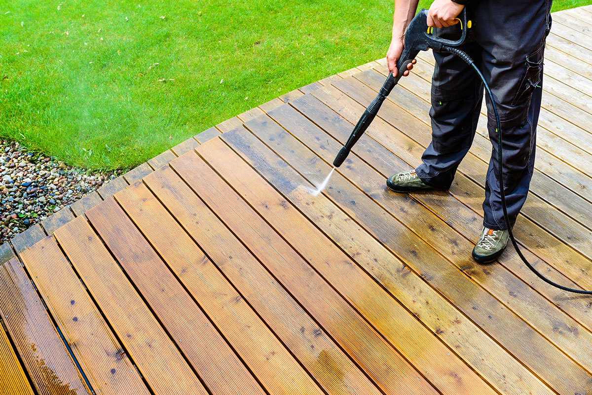 A person uses a pressure washer to clean a wooden deck, revealing a striking difference between the washed and unwashed sections. Dressed in dark pants and sturdy shoes, they showcase the power of a pressure washing service in Pike County KY. In the background, a lush lawn spreads out vividly.