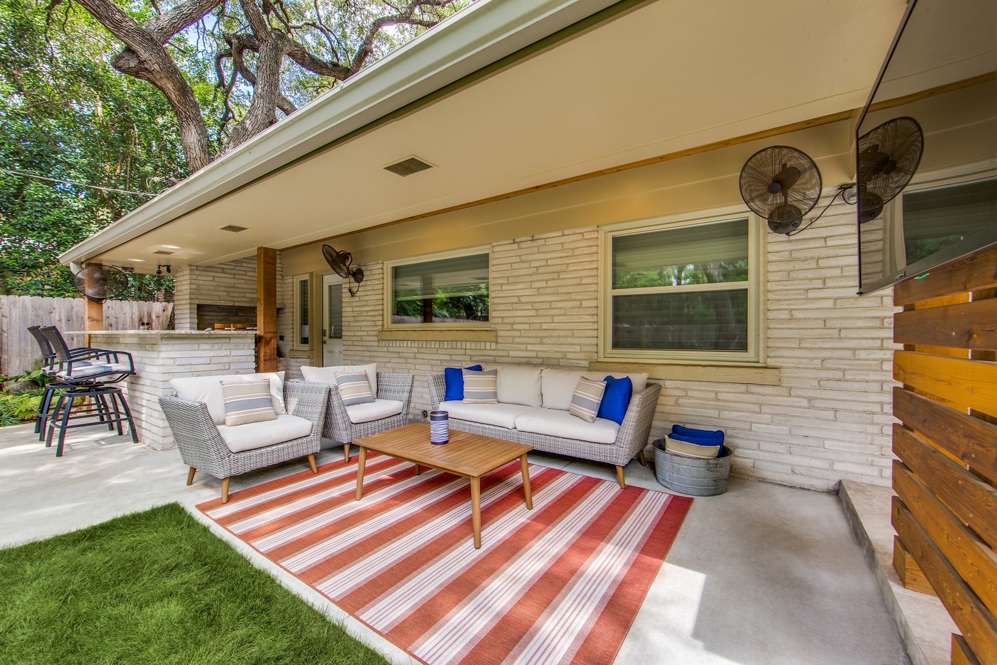 Outdoor patio with a striped red and white rug, wicker sofas with white cushions, and blue throw pillows. A wooden coffee table is centered. Bar stools are by a counter in the background. Ceiling fans and a wooden privacy screen complete the cozy setting, freshly cleaned by Pike County KY's pressure washing service.