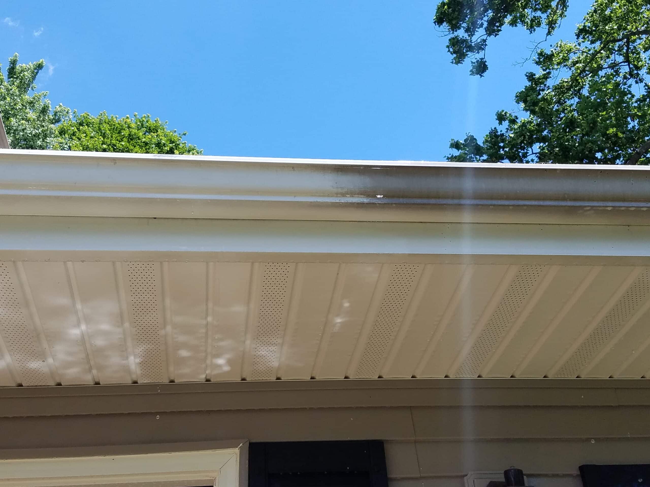 White house eaves with a metal gutter are shown against a blue sky, hinting at the expertise of the best roof washing service in Northeast Kentucky. The gutter has a section with dark staining, while trees with green leaves are visible in the background.