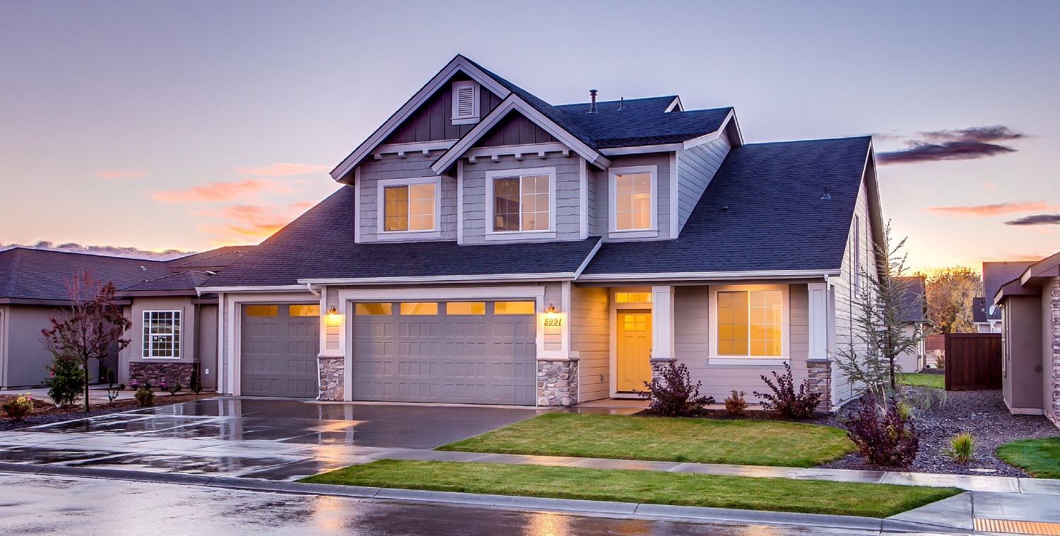 A modern two-story house with a gray exterior and two garages, illuminated by sunset light, shines after a top-notch pressure washing service in Cabell County, WV. The wet driveway and lawn suggest recent rain. The sky’s orange and purple hues enhance the serene suburban setting.