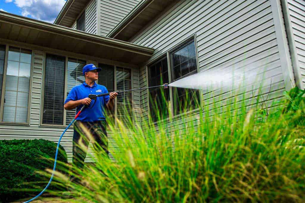 A person in a blue shirt and cap is power washing the siding of a beige house, using a high-pressure spray to blast away dirt. Surrounded by lush plants and grass, it’s the ultimate showcase of pressure washing service in Pike County, KY.