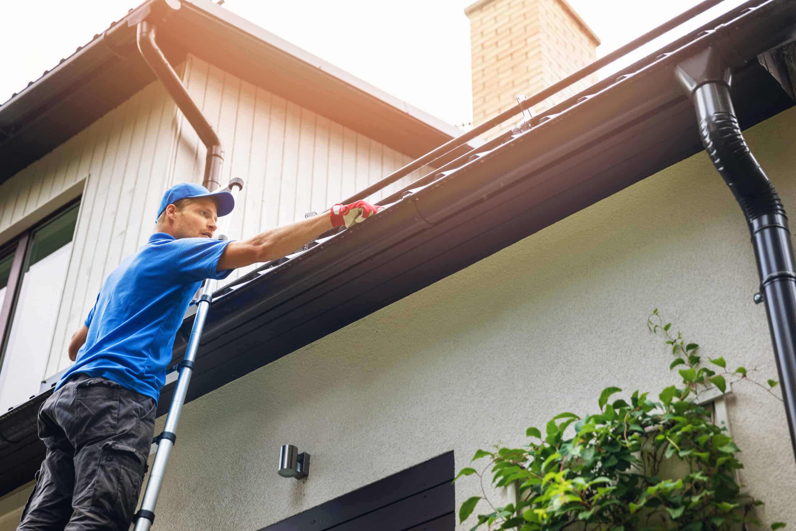 A person in a blue shirt and cap stands on a ladder cleaning a house's gutter with the precision of the best window cleaning service in Northeast Kentucky. Using a gloved hand to remove debris, they work under the bright sun on a house with beige siding, a chimney, and lush greenery below.