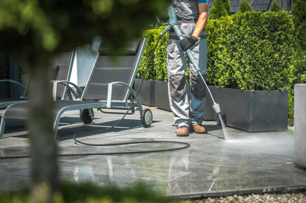 A person from a pressure washing service in Fayette County, KY, meticulously cleans a patio with large gray tiles surrounded by potted shrubs. Dressed in work attire and gloves, they focus on cleaning the stone surface with a water jet, while two patio chairs stand nearby.