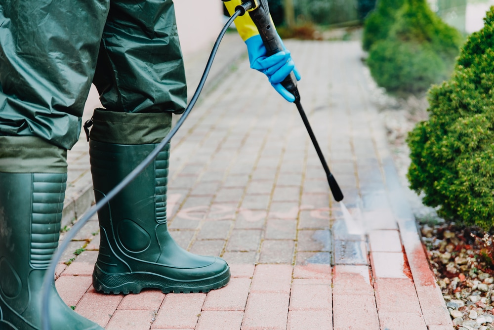 A person in green protective clothing and rubber boots expertly operates a pressure washer to clean a brick walkway. Blue gloves are noticeable as water sprays out, creating a vivid splash. This is the quality you can expect from the top pressure washing service in Northeast Kentucky.