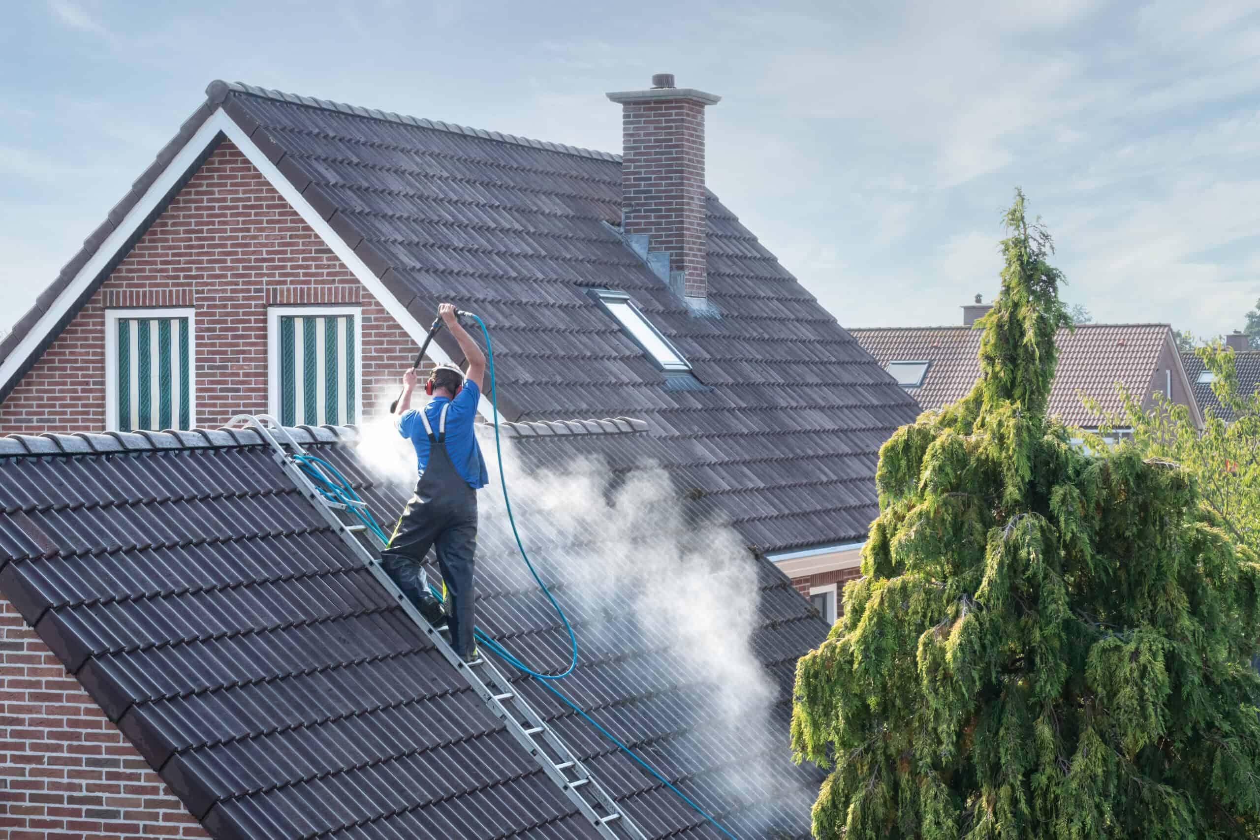 A person in blue overalls is power washing the roof of a brick house, standing on a ladder. Surrounded by mist and dark shingles, they expertly handle the job near a tall, green tree. This scene could be straight out of the best roof washing service Northeast Kentucky has to offer.