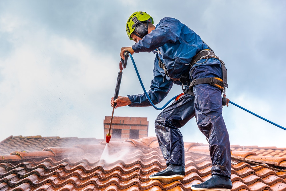 A worker in a helmet and protective gear is power washing a red tiled roof under cloudy skies. Using a high-pressure hose, the worker is safely secured with a harness, showcasing the best roof washing service in Northeast Kentucky.