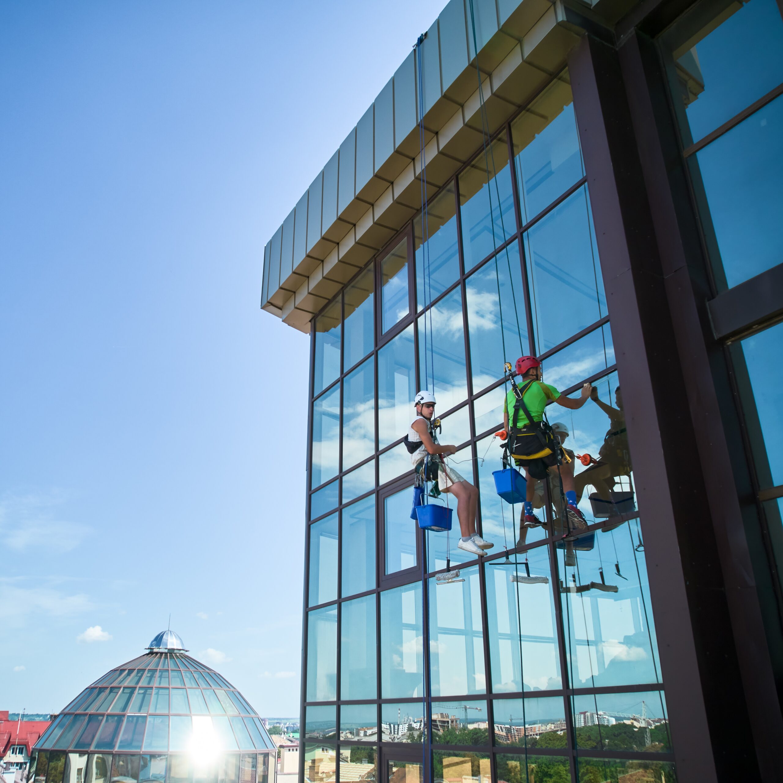 Two workers in safety gear expertly clean tall glass windows of a modern building, suspended on ropes with buckets in hand. Under the clear sky and with a dome structure in the background, they exemplify the precision of our pressure washing service in Northeast Kentucky.