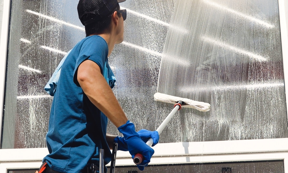 A person wearing a black cap, blue shirt, and gloves is cleaning a large, glass window with a squeegee. The window gleams as light reflects off the soapy water. This scene captures the commitment to excellence you'd expect from the best roof washing service in Northeast Kentucky.