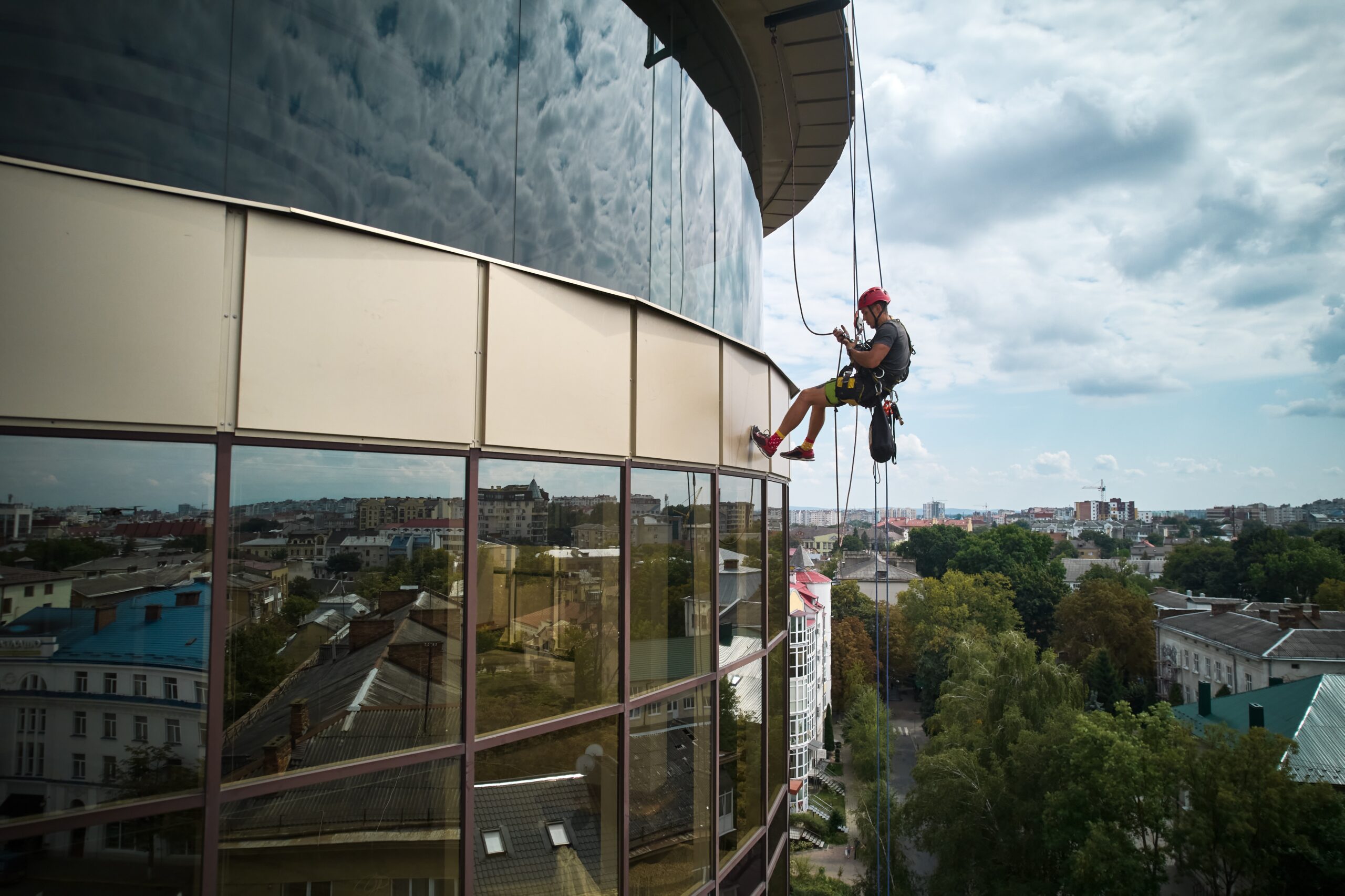 A worker in safety gear hangs from ropes while offering the best window cleaning service in Northeast Kentucky on a tall building. The cityscape reflects off the glass facade. Amid cloudy skies, trees and urban structures form a picturesque backdrop for this high-altitude task.