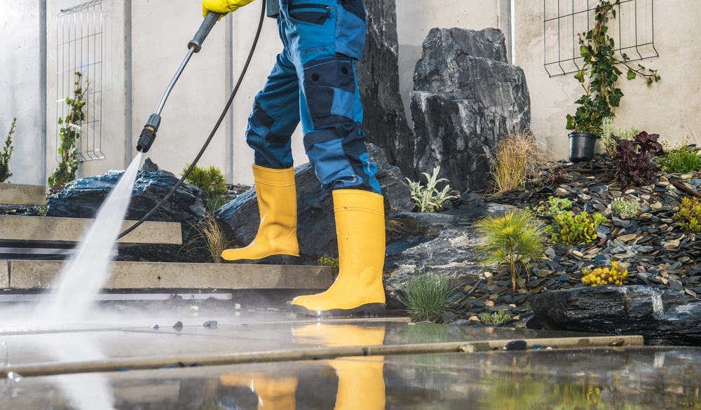 A person wearing yellow boots and blue overalls is pressure washing a patio in Pike County, KY. Water sprays from the hose, cleaning the stone surface. Decorative rocks and plants are in the background, showcasing a professional touch unique to the best services in Northeast Kentucky.