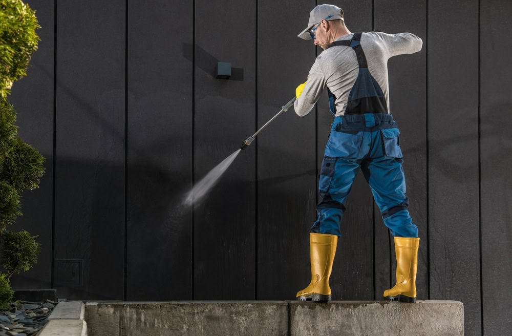 A person wearing a gray shirt, blue overalls, and yellow rubber boots is power washing a dark wall, showcasing the efficiency of our pressure washing service in Fayette County, KY. Holding a precision nozzle, they spray water on the wall as a small shrub peeks into view on the left.