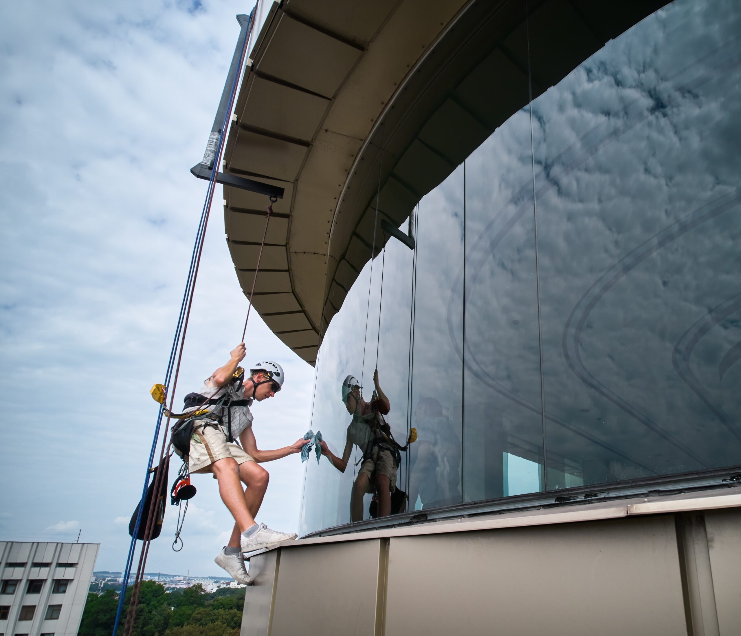 A window cleaner in a helmet and safety harness dangles from a rope, expertly cleaning the large curved glass windows on a towering building. The cloudy sky and distant structures frame his work, reminiscent of pressure washing service professionals in Pike County KY.