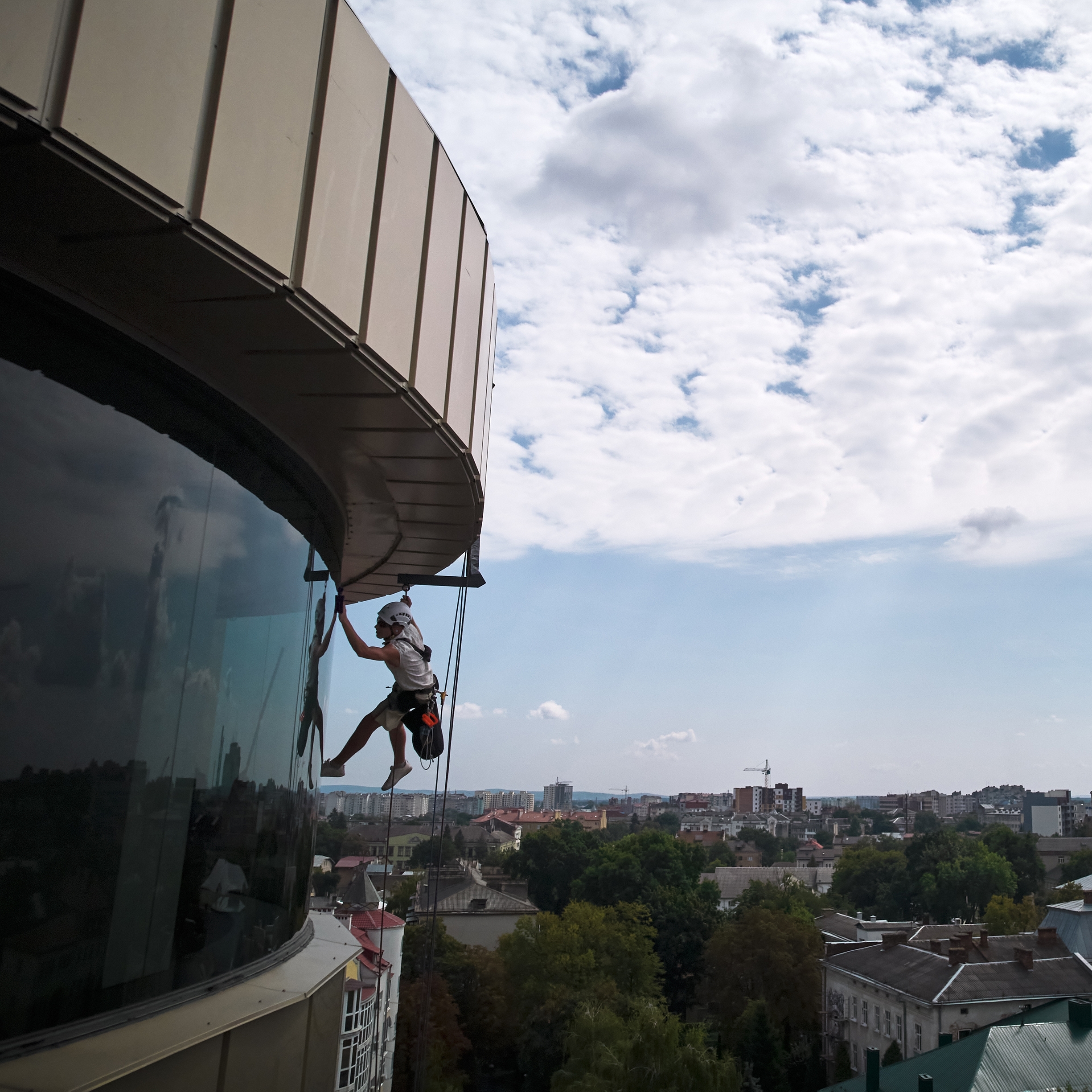 A window cleaner suspended by ropes works on the glass exterior of a tall building, akin to the precision of the best roof washing service in Northeast Kentucky. A cityscape with scattered clouds is visible in the background, framing their meticulous work.