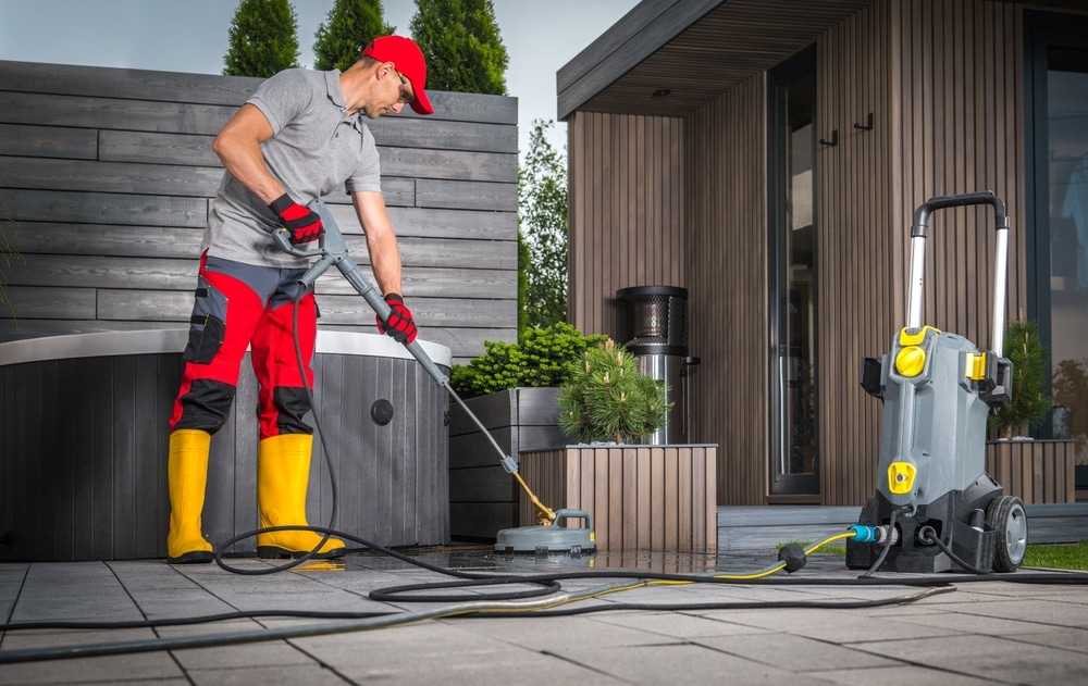 A person in a red cap, gray shirt, red and black pants, and yellow boots is using a power washer to clean a patio. Nearby, the power washing service Northeast Kentucky machine hums efficiently. In the background, a modern building and potted plants complete the scene.