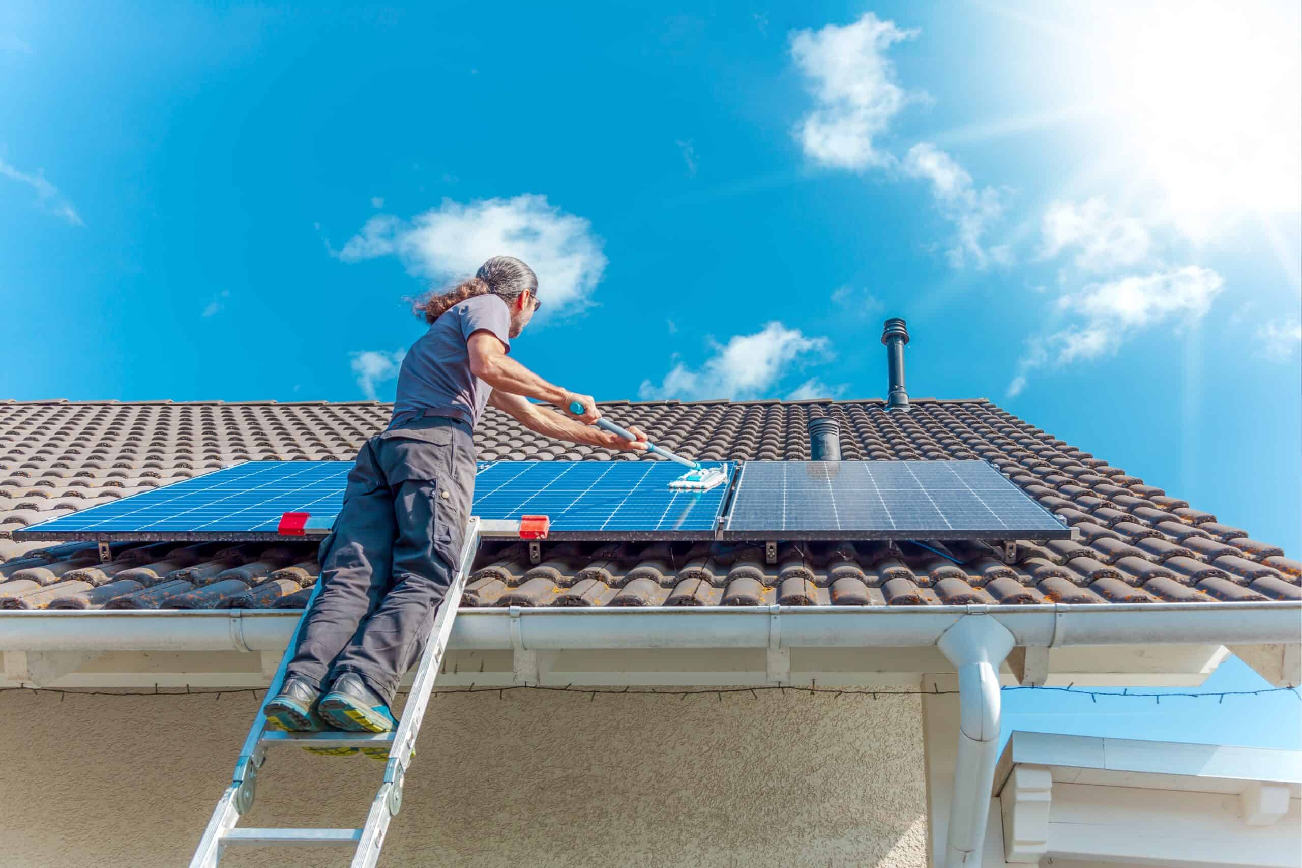 A person standing on a ladder cleaning solar panels on a sloped roof under a bright blue sky with a few clouds. The sun is shining, casting reflections on the panels. The person, possibly from the best window cleaning service in Northeast Kentucky, wears a gray shirt and black pants.