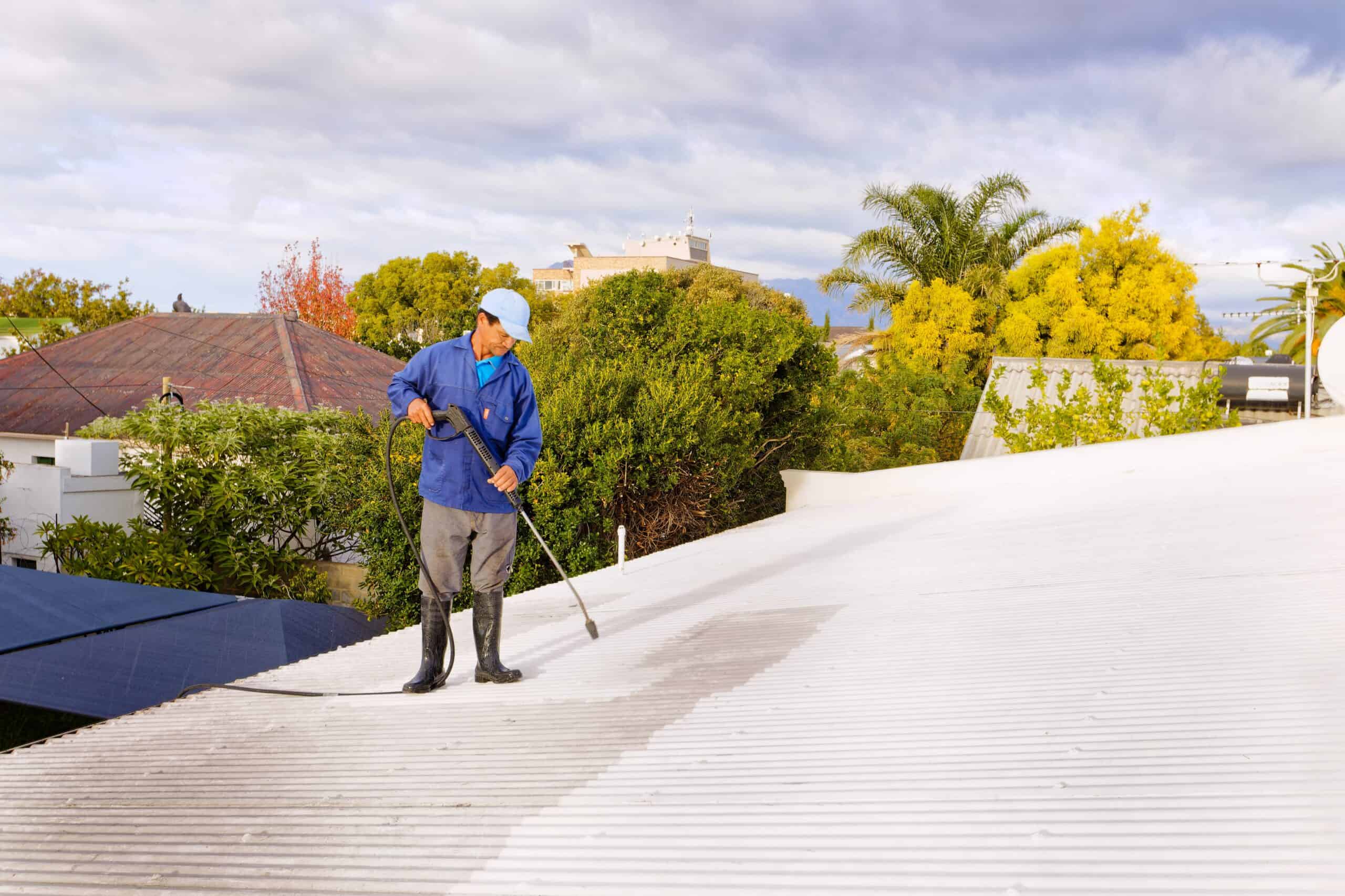 A person in a blue shirt, cap, and boots uses a pressure washer to clean a white corrugated roof. Engaged in providing pressure washing service across Northeast Kentucky, they are surrounded by trees and neighboring houses under a partly cloudy sky.