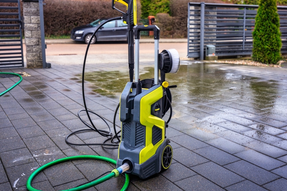 A yellow and black pressure washer sits on the wet, paved driveway of a car near a gate. With wheels, a green water hose, and a white brush on top, it's ready for a pressure washing service in Pike County KY or Northeast Kentucky.