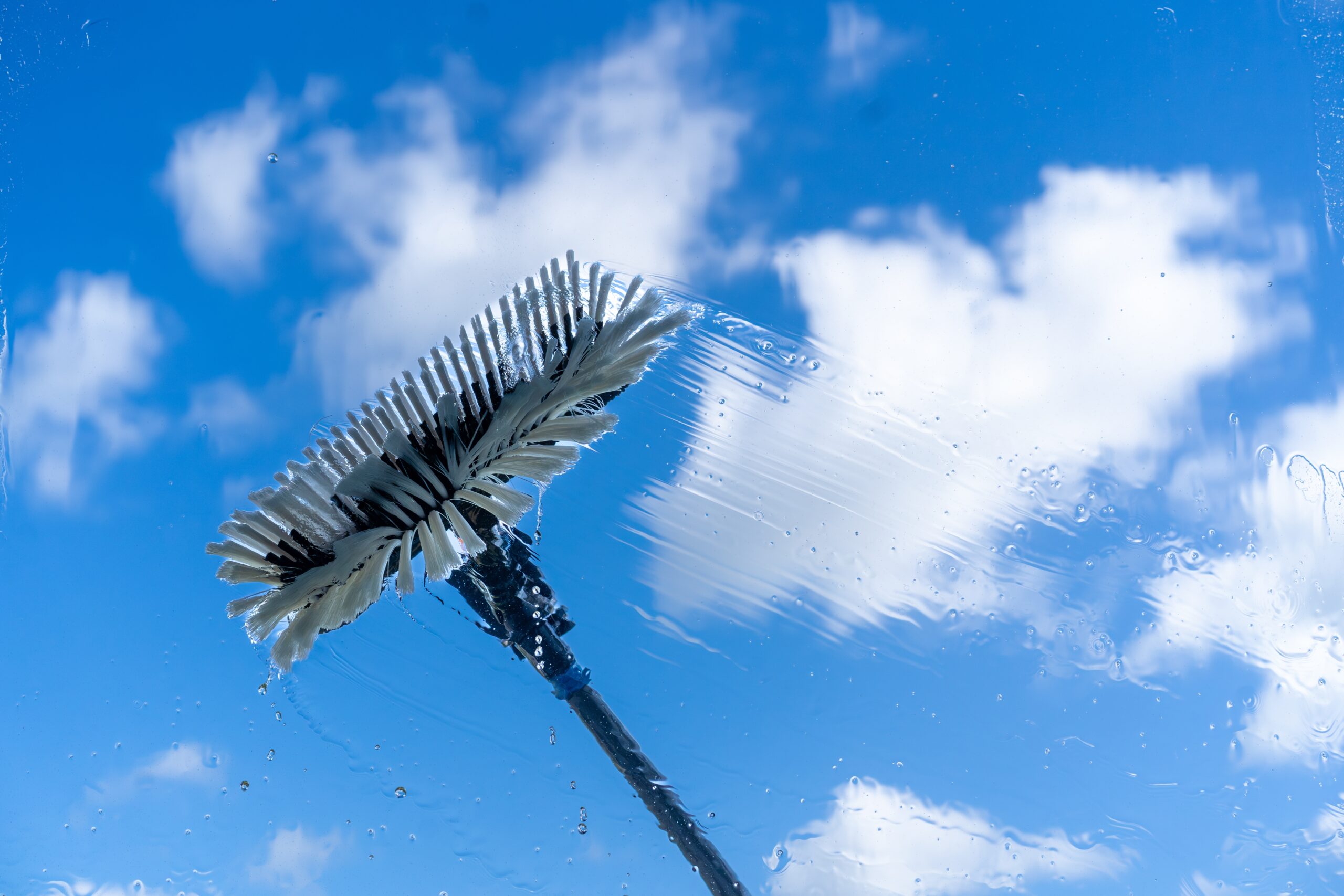 A window cleaning brush swipes across the glass against a bright blue sky dotted with clouds. This mid-swipe action showcases the best window cleaning service in Northeast Kentucky, leaving clear paths among droplets and streaks, promising crystal-clear windows every time.