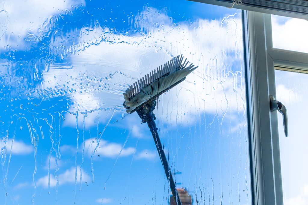 A person is cleaning a large window using a brush with an extendable pole, reminiscent of a professional pressure washing service in Fayette County, KY. The clear blue sky with scattered white clouds is visible through the glass, along with water streaks and soap suds on the window's surface.