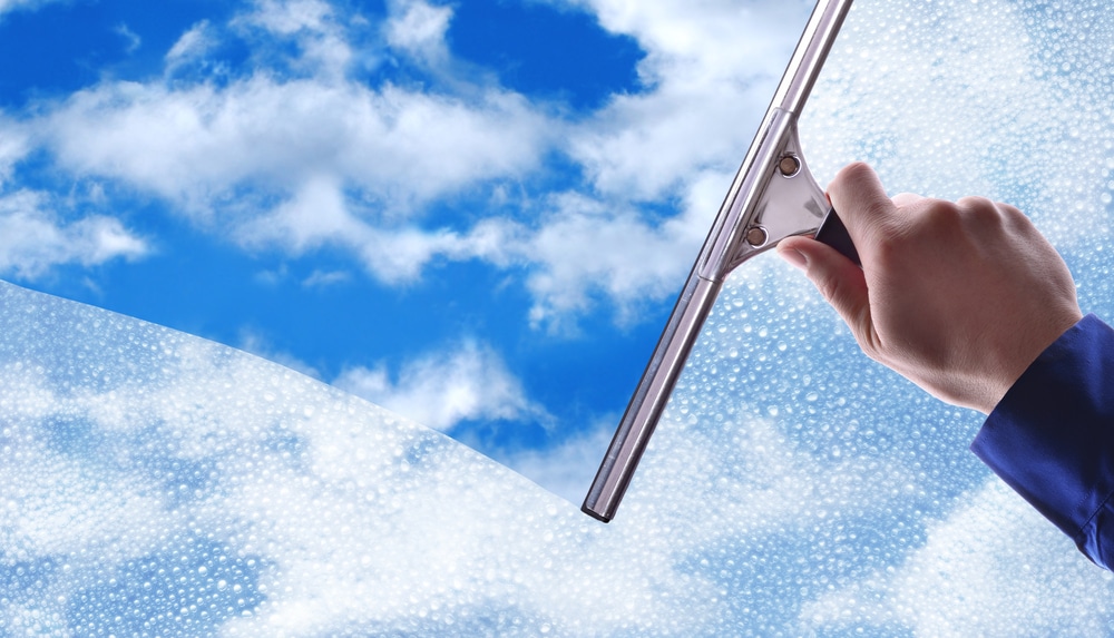 A hand using a squeegee to clean a window, revealing a clear view of blue sky with fluffy white clouds. Soap suds linger on the part yet to be cleaned, showcasing the pristine results you can expect from our pressure washing service in Northeast Kentucky.